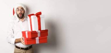 Kış tatili ve kutlama. Heyecanlı adam elinde Noel hediyeleri tutuyor ve şaşırmış görünüyor, Noel Baba şapkası takıyor, beyaz arka planda duruyor..
