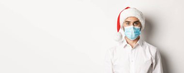 Covid-19 kavramı, sosyal mesafeler ve kış tatilleri. Coronavirus 'tan Noel Baba şapkası ve yüz maskesi takan genç adamın yakın çekimi. Yeni yılı kutluyoruz..