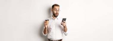 Kahve içen ve cep telefonundaki mesaja şaşıran bir iş adamı beyaz arka planda şaşkın şaşkın dikiliyor..