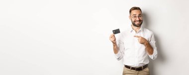 Gözlüklü yakışıklı adam kredi kartını işaret ediyor, banka hizmetlerinden memnun, beyaz arka planda duruyor..