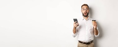 Yakışıklı adam kahve içmeyi başarıyor, cep telefonundan gelen mesaja şaşırıyor, beyaz arka planda duruyor..