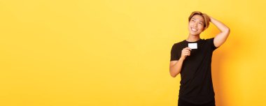 Düşünceli gülümseyen Asyalı adam kredi kartı gösterirken düşünüyor, sol üst köşeye bakıyor rüya gibi, sarı arka plan.