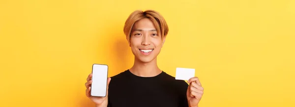 スマートフォンの画面とクレジットカードを示す笑顔ハンサムなアジアの男のクローズアップ 立って黄色の背景 — ストック写真