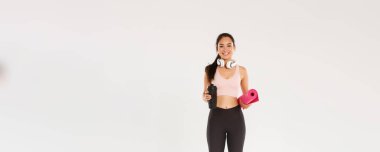 Yoga derslerine giden Asyalı güzel bir kız, kulaklıklı ve su şişeli bir bayan sporcu spor salonu eğitimi için lastik paspas taşırken gülümsüyor..
