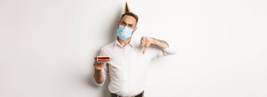 Covid-19, sosyal uzaklık ve kutlama. Yüz maskesi takan hayal kırıklığına uğramış doğum günü çocuğu, elinde dilek mumuyla doğum günü pastası tutuyor, hoşlanmadığını ifade etmek için başparmağını aşağı indiriyor, beyaz arka plan.