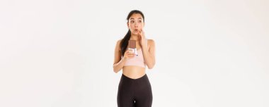 Spor, refah ve aktif yaşam tarzı konsepti. Düşünceli ve kararsız Asyalı kız kalori ve diyet düşünüyor, spor yapıyor, çikolatayı tutuyor, yemeye çekiniyor, beyaz arka plan.