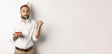 Tatiller ve kutlamalar. Neşeli adam doğum gününün tadını çıkarıyor, parti düdüğü çalıyor ve elinde doğum günü pastası tutuyor, beyaz arka plan.