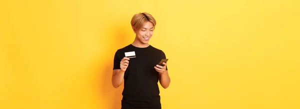 オンライン購入 インターネットでの買い物 スマートフォンやクレジットカードの保有 黄色の背景のために支払うハンサムなアジアの男を笑顔 — ストック写真