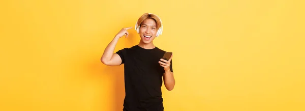 满意的亚洲人喜欢音乐或播客 用手指指著耳机 拿着智能手机 站在黄色的背景上 — 图库照片