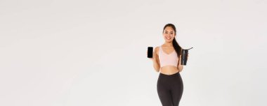 Tam boy gülümseyen Asyalı fitness kızı, aktif giyimli spor kadın akıllı telefon ekranı ve su şişesini gösteriyor, eğitim sırasında mobil uygulama ile içme sıvısını kontrol ediyor..