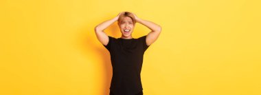 Genç Asyalı adamın portresi panikliyor, endişeli ve üzgün görünüyor, el ele tutuşuyor ve yüzünü buruşturuyor, sarı arka planda duruyor..