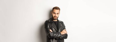 Çekici, sakallı, deri ceketli, gülümseyen, elleri göğsünde çaprazlanmış, beyaz arka planı olan maço bir adam..