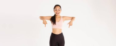 Spor, refah ve aktif yaşam tarzı konsepti. Heyecanlı ve mutlu Asyalı spor giyim kızı, parmakları aşağıda ve büyük indirimlerle şaşkın bakıyor, afiş okuyor, beyaz arka plan.