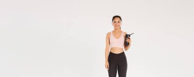 Tam boy gülümseyen ince ve mutlu fitness kızı, spor salonunda kulaklıkla çalışan Asyalı bayan sporcu, antrenman sırasında müzik dinleyen, egzersizler arasında şişeden su içen, beyaz arka plan.