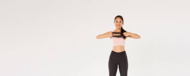 Tam boy ince ve fit görünümlü Asyalı kız spor yapıyor, direnç bandını elleriyle geriyor ve gülümsüyor. Spor salonunda egzersiz malzemesi kullanan antrenör, beyaz arka plan..