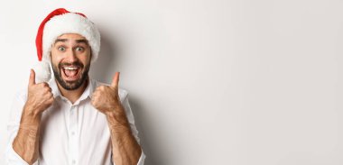 Parti, kış tatili ve kutlama konsepti. Noel 'in tadını çıkaran, Noel Baba şapkası takan ve heyecanlı yüzüyle baş parmağını gösteren beyaz bir adam..