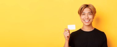 Sarışın, mutlu ve sarı arka planda kredi kartı gösteren yakışıklı Asyalı adamın yakın çekimi..