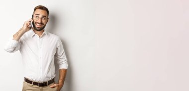 Gözlüklü başarılı bir iş adamı cep telefonuyla konuşuyor, tatmin olmuş görünüyor ve gülümsüyor, beyaz arka plana karşı duruyor..