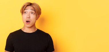 Etkilenmiş Asyalı yakışıklının yakın çekimi, sol üst köşesine bakıyor, çenesi düşmüş ve şaşırmış, sarı arka planda duruyor..