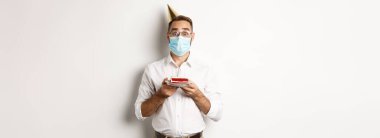 Covid-19, sosyal uzaklık ve kutlama. Sürpriz doğum günü pastası tutan çocuk, Coronavirus 'un yüz maskesi takıyor, beyaz arka plan.