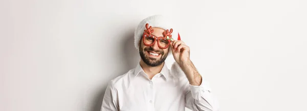 stock image Close-up of handsome bearded guy in xmas party glasses and santa hat, smiling and wishing merry christmas, standing over white background.