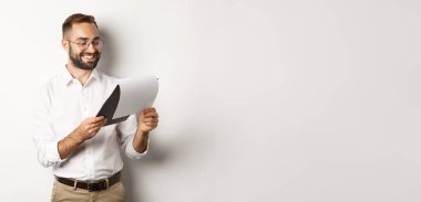 İşadamı belgelerden memnun görünüyor, rapor okuyor ve gülümsüyor, beyaz arka planda duruyor..