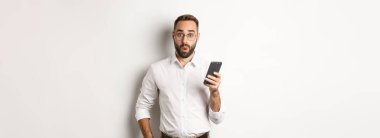 Şaşkın yakışıklı adam internette ilginç bilgiler okuyor, elinde akıllı telefon, beyaz arka planda duruyor..