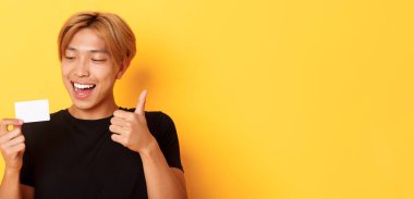 Mutlu ve memnun Asyalı yakışıklı adamın yakın plan fotoğrafı, kredi kartı ve onay kartı gösteriyor, şaşırmış bir şekilde gülümsüyor, sarı arka planda duruyor.