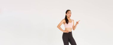 Spor ve spor konsepti. Tam boy şaşırmış ve şaşırmış, spor aletleri ya da spor salonu üyelik fiyatları gösteren, üst sol köşedeki işaret eden ve bakan, güzel Asyalı kız..