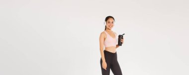 Tam boy gülümseyen ince ve mutlu fitness kızı, spor salonunda kulaklıkla çalışan Asyalı bayan sporcu, antrenman sırasında müzik dinleyen, egzersizler arasında şişeden su içen, beyaz arka plan.