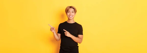 満足した笑顔ブロンドの男の肖像画 アジアの学生の指を指して右 方法を示す 立って黄色の背景 — ストック写真