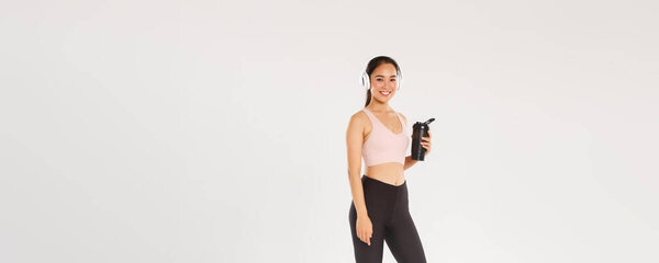 Полная длина улыбается стройная и счастливая девушка фитнес, азиатская женщина athelte тренировки в тренажерном зале с наушниками, прослушивание музыки во время тренировки, питьевая вода из бутылки между упражнениями, белый фон.
