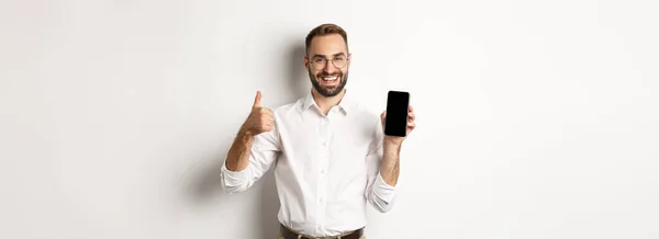 满意的商人戴着眼镜 竖起大拇指 演示手机屏幕 推荐应用程序 站在白色背景之上 — 图库照片