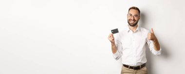 Yakışıklı bir adam kredi kartını ve başparmağını kaldırmış, bankayı tavsiye ediyor, beyaz arka planda duruyor. Boşluğu kopyala