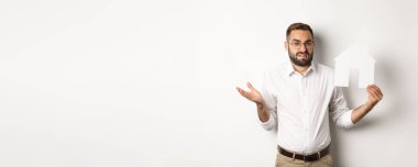 Gayrimenkul. Kafası karışmış bir adam omuz silkiyor, ev kâğıdı modelini gösteriyor ve kararsız görünüyor, beyaz arka planda duruyor. Boşluğu kopyala