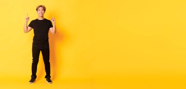 Uzun boylu, kendine güvenen yakışıklı, sarı saçlı, gülümseyen ve parmakla işaret eden, logo gösteren, sarı arka planlı Asyalı bir adam..