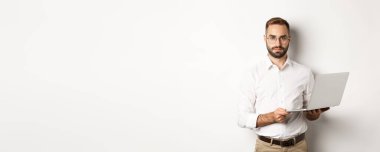 İş için. Bilgisayarda çalışan yakışıklı bir yönetici, elinde bilgisayarla kameraya bakıyor, beyaz arka planda duruyor..