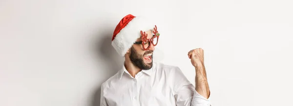 快乐的大胡子男人戴着圣诞礼帽 戴着圣诞眼镜 做着拳头泵 兴高采烈 达到目标 庆祝一番 白色背景 — 图库照片