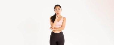 Spor, refah ve aktif yaşam tarzı konsepti. Düşünceli ince ve formda Asyalı fitness kızı spor salonunu seçiyor, düşünüyor ya da karar veriyor, kameraya bakıp eğleniyor ve aktif giysinin içinde duruyor..