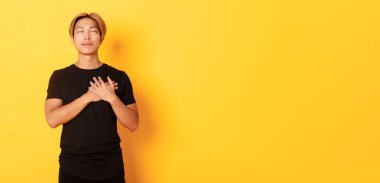 Dokunulmuş yakışıklı Asyalı adamın portresi. Kalbinin üstünde el ele tutuşmuş, gururu okşanmış, sarı arka plan..