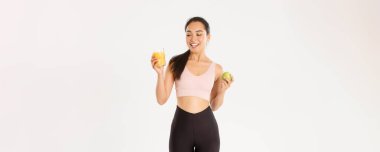 Spor, refah ve aktif yaşam tarzı konsepti. Spor giyim kuşağında gülümseyen Asyalı fitness kızı portakal suyuna bakıyor, spor salonunda egzersiz yaptıktan sonra elma yiyor, beyaz arka plan..