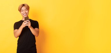 Sarı saçlı yakışıklı Asyalı adamın portresi, cep telefonu kullanıyor ve gülümsüyor, akıllı telefon uygulamasında mesajlaşıyor, sarı arka plan.