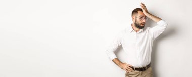 Sinirli adam yönetici gözlerini yuvarlar ve alnını tokatlar. Yorucu bir şeyin yüzü, beyaz arka planda durur..