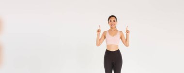 Spor, spor salonu ve sağlıklı vücut konsepti. Neşeli bir gülümseme ve hoş bir fitness kızı, parmaklarını havaya kaldırarak egzersiz malzemeleri reklamını gösteriyor, eğitim için üyelik satın alıyor..