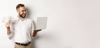 İş ve e-ticaret. Tatmin olmuş iş adamı dizüstü bilgisayarda iş yapıyor ve elinde para tutuyor, mutlu bir şekilde gülümsüyor, beyaz arka planda duruyor..