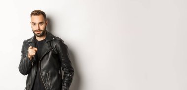 Yakışıklı ve havalı sakallı adam kamerayı işaret ediyor, deri ceket giyiyor, beyaz arka plana karşı küstah duruyor..