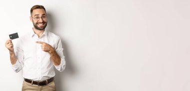 Gözlüklü yakışıklı adam kredi kartını işaret ediyor, banka hizmetlerinden memnun, beyaz arka planda duruyor..