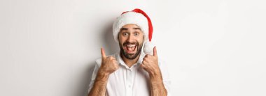 Parti, kış tatili ve kutlama konsepti. Noel 'in tadını çıkaran, Noel Baba şapkası takan ve heyecanlı yüzüyle baş parmağını gösteren beyaz bir adam..