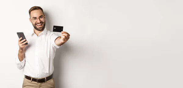 ビジネスとオンライン決済 スマートフォンを持ちながらクレジットカードを見せる興奮した男は白い背景に満足して立っていた — ストック写真