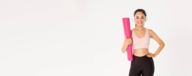 Spor, refah ve aktif yaşam tarzı konsepti. Şımarık, güzel Asyalı fitness kızı, yoga derslerine ya da ev eğitimine hazır bir kadın, antrenman için lastik minder tutuyor ve kulaklıkla müzik dinliyor..